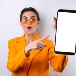 young-woman-trendy-stylish-glasses-bright-orange-oversized-jacket-white-background-holds-phone-with-blank-white-screen